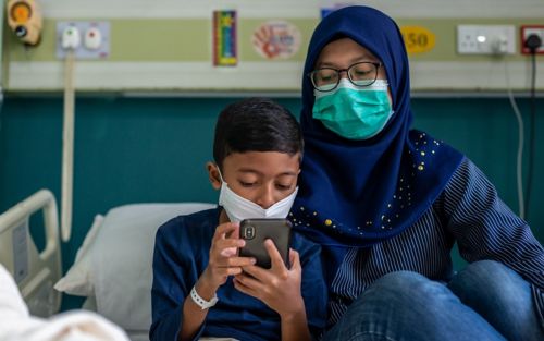 Boy and mother wearing face masks, looking at phone while sitting on a hospital bed.