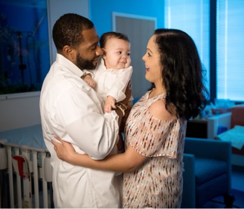 अस्पताल के कमरे में माता-पिता अपने बच्चे का हाथ पकड़कर खड़े हैं।