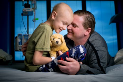 Young cancer patient on a hospital bed smiling, holding a stuffed dog toy, and touching foreheads with their father, who is kneeling on the floor and leaning on the bed.