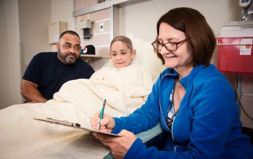 Trabajadora social de oncología pediátrica completando formularios junto a un padre y un joven paciente en la cama del hospital.
