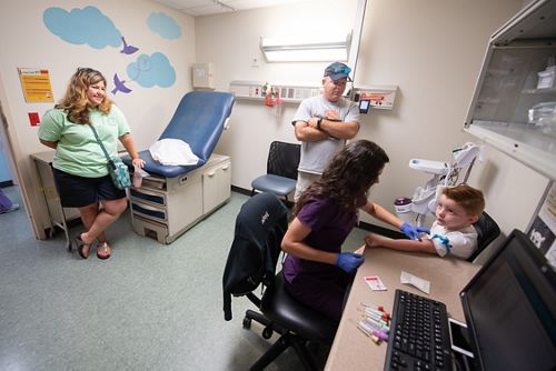 Male child patient receives blood draw from female nurse in purple scrubs with parents in background.