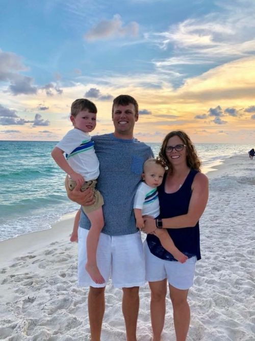 A family standing on the beach in front of a sunset, with two parents holding two children.