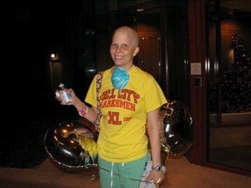 A patient standing in a yellow shirt, holding balloons and a drink bottle, with a blue mask draped around their neck.