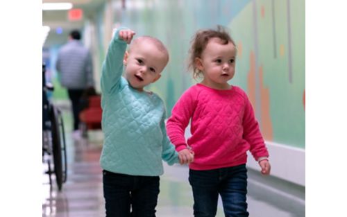 Two children hold hands while walking down hospital hallway