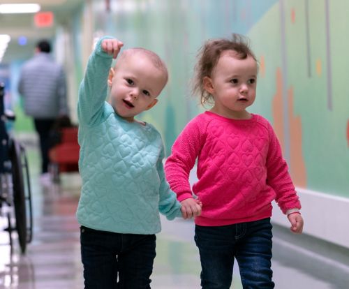 Two children hold hands while walking down hallway at hospital