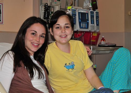 Patient laying in bed in a white shirt with her sister standing next to the bed and leaning down.