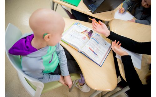 Child cancer patient sitting at school desk with open textbook.