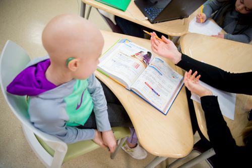 Pediatric cancer patient sits in desk with text book open in conversation with unseen teacher.
