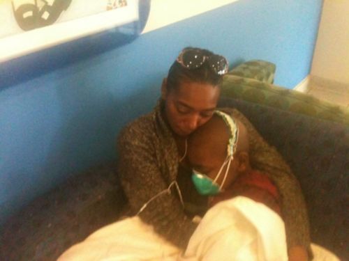 A young cancer patient sitting on a couch receiving a hug from his mom.