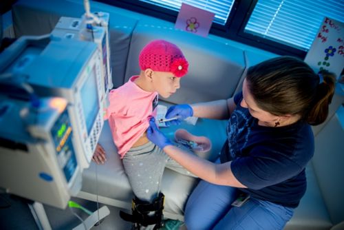 A young cancer patient sitting on a couch receives medicines through a port from a nurse.