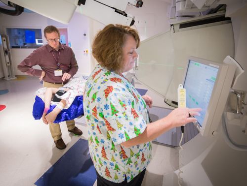 Radiation therapist prepares the computer for a pediatric cancer radiation treatment with another radiation therapist and a patient in the background.