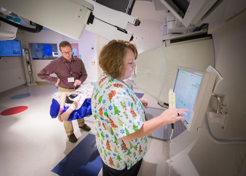 Radiation therapist prepares the computer for a pediatric cancer radiation treatment with another radiation therapist and a patient in the background.