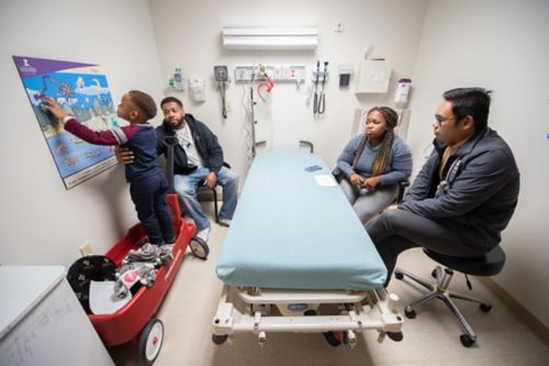 On top of daily needs and life demands, families facing childhood cancer also have to talk and plan with the medical team and learn about medicines and medical care. In this image, a family meets with a physician in a clinic room. The father keeps the young patient from falling off of a wagon as the doctor reviews results.