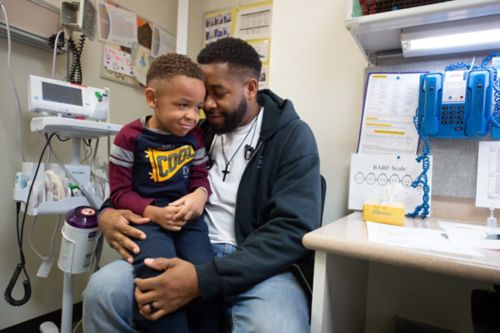 Responda a las preguntas acerca del dolor con honestidad. En esta foto, un paciente pequeño con cáncer y su papá están sentados juntos y, detrás de ellos, hay una escala BARF.