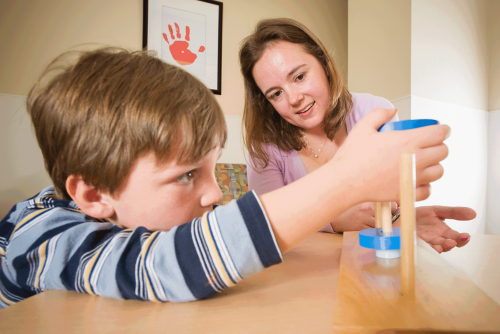 Childhood cancer survivor participates in a neuropsychological assessment with a psychologist. He is leaning over a table trying to balance a wooden block onto a dowel. 