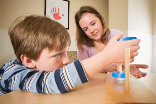 O sobrevivente de câncer infantil participa de uma avaliação neuropsicológica com um psicólogo. Ele está se inclinando sobre uma mesa tentando equilibrar um bloco de madeira em uma cavilha.