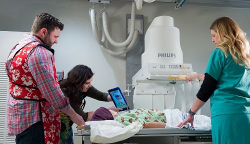Ребенок с онкологическим заболеванием лежит на столе, в то время как специалист по адаптации ребенка показывает ему iPad, а рентгенотехник размещает рентгеновский аппарат для рентгенографии брюшной полости.
