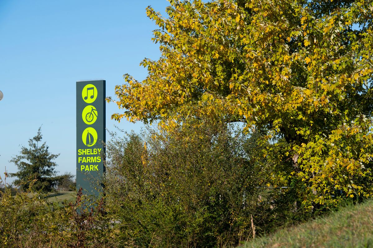 Trail sign in a bush with a music note, bike, and boat icon that reads Shelby Farms Park.