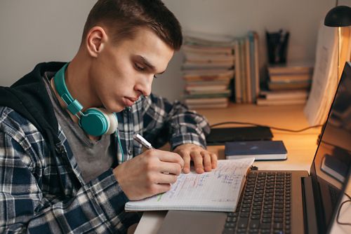 A student wearing headphones sitting and a computer and writing on a notebook
