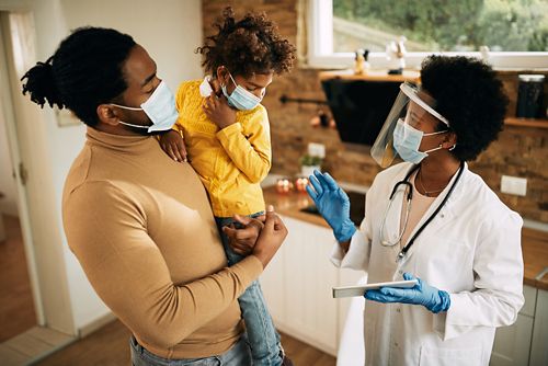 Father holds young son while getting home vaccine from doctor in shield face mask