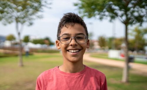 A portrait of a young boy with glasses smiling outdoors in front of trees.
