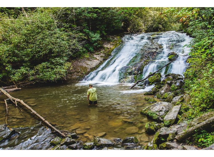 Man standing in the water fishing in front of a waterfall.