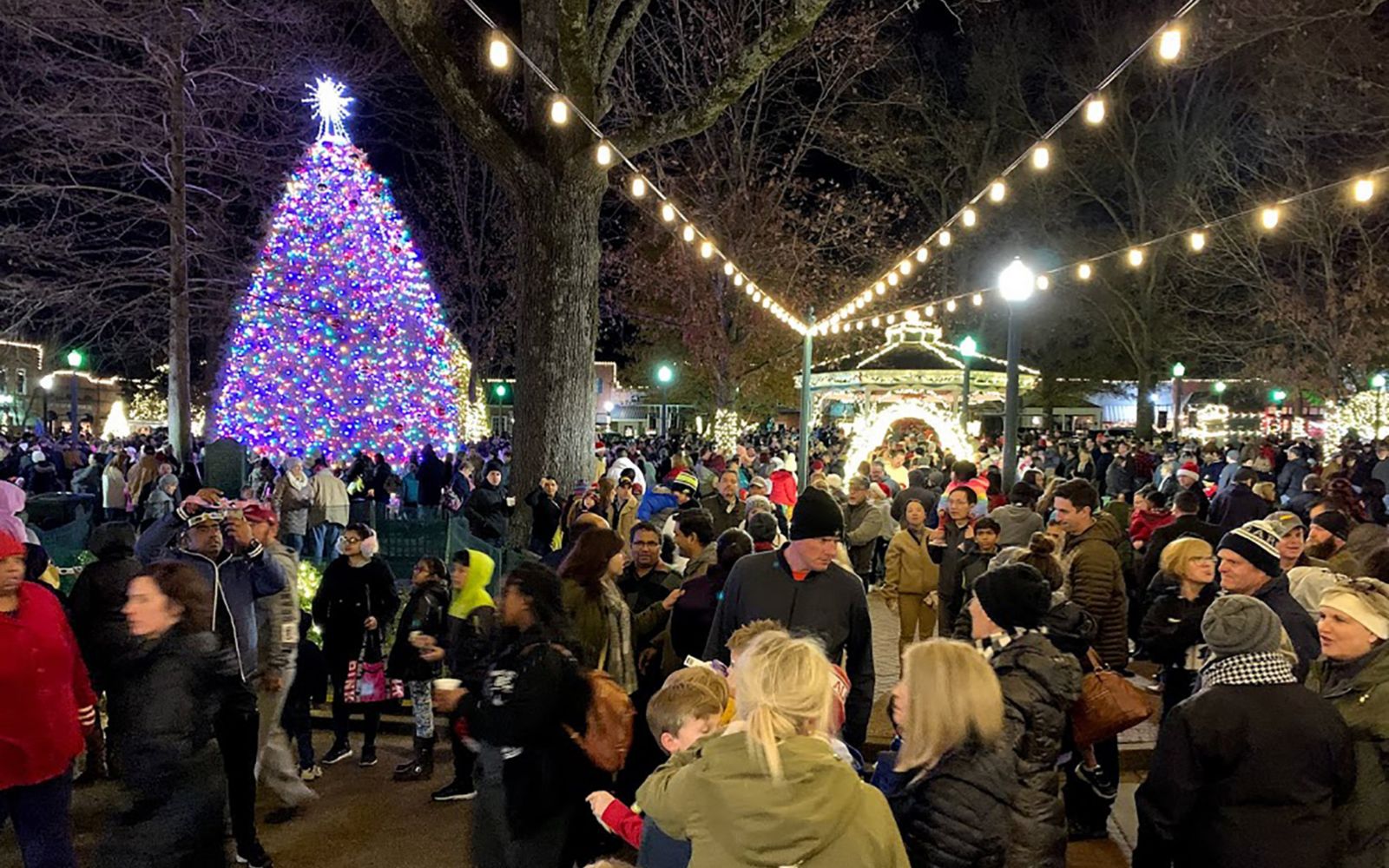 A crowd of people in a town square around a lit Christmas tree at night.