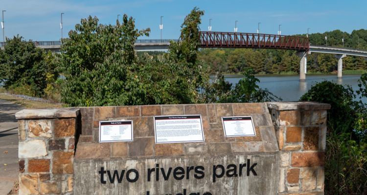 Plaque that reads "Two Rivers Park Bridge" with a bridge over the river in the background.