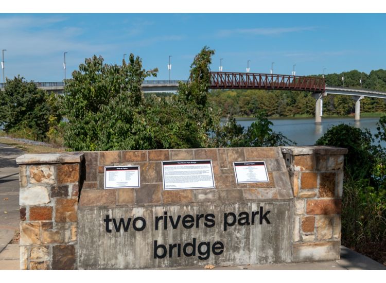 Plaque that reads "Two Rivers Park Bridge" with a bridge over the river in the background.