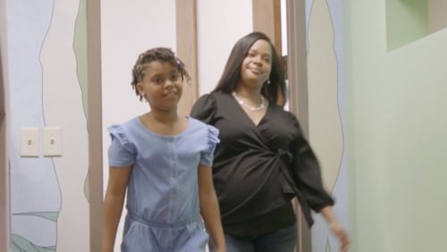 Mother and daughter walking together in medical clinic hallway, young girl in blue dress with woman in black outfit, health care facility interior