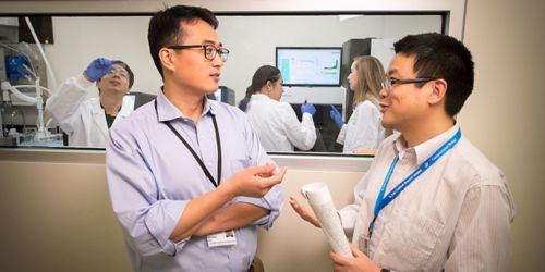 Two men talking outside a lab where people are working