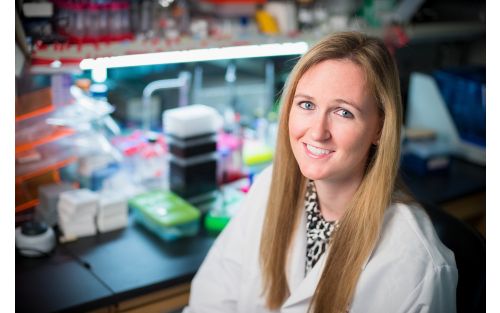 Researcher in lab coat sitting in a lab and smiling at camera.