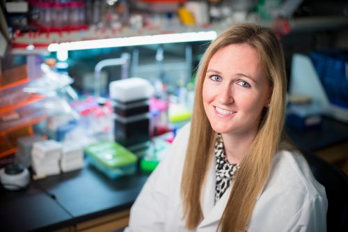 Researcher in lab coat sitting in a lab and smiling at camera.