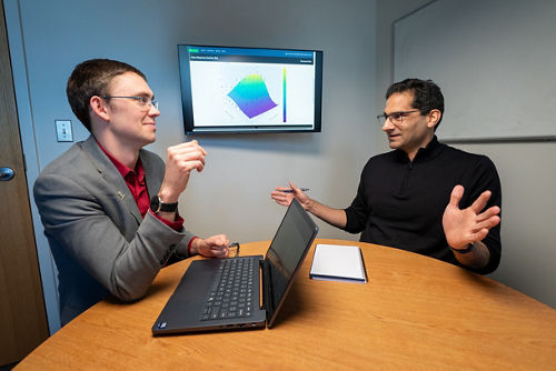 Two men sitting at a table with a computer having a conversation