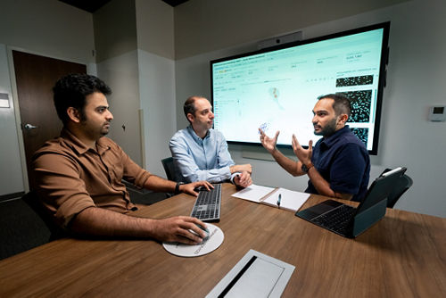 Three men at a table with a computer screen on the wall.