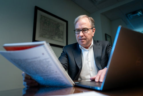 A man in a gray suit reads a notebook with his hand on a computer