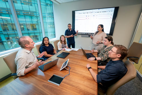 Meeting in conference type room. People around table, 2 presenting at screen.