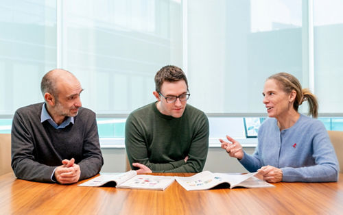 Two men and a woman seated at a table, talking about something in a magazine