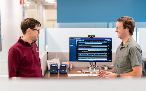 two men talking in front of a double screen computer monitor