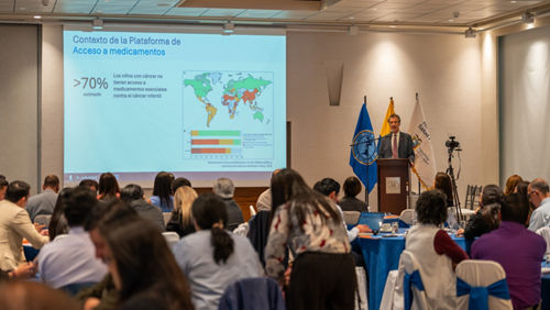 Man at podium speaking to group seated at tables in front of a large screen showing a world map