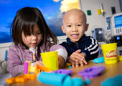 Pediatric cancer patient plays with modeling clay in a hospital bed with his sister.