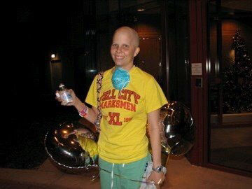 A patient standing in a yellow shirt, holding balloons and a drink bottle, with a blue mask draped around their neck.